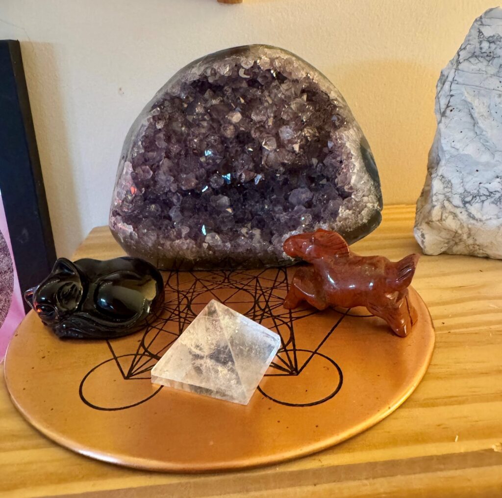 A copper crystal grid on a wooden shelf in my healing room, with an amethyst geode, an obsidian cat, a red jasper horse and a clear quartz pyramid.