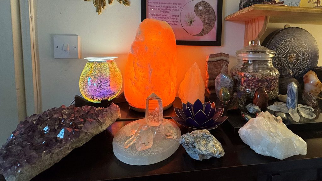A display of crystals in my healing room, including an heart shaped amethyst cluster, a glowing salt lamp, a geometric oil burner, a selenite plate with clear quart points, a lotus candle holder, a piece of Kyanite, a Lemurian quartz piece and a clear quartz cluster.